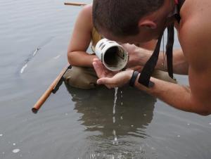 a person wearing waders stands and water and pours something from a plastic sample cup into their hand. A second person crouches in the water nearby.